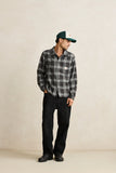 Man wearing a plaid shirt, black pants, and a cap against a beige background Rhythm Alex Check LS Shirt - Forest
