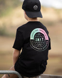 A child wearing a black t-shirt and black cap, viewed from behind, sitting on a wooden fence. The back of the t-shirt features a large circular graphic with the text 'CREATE THE IMPOSSIBLE' in a light green-to-pink gradient, 'UNIT' in the center, and 'PERFORMANCE DIVISION' in white. Unit Youth Tees - Evolve - Black