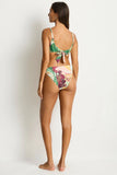 A woman modeling a two-piece bikini from the back, featuring a green and white patterned top with a tie-back knot and matching high-cut bottoms with a multicolor abstract print, set against a plain white studio background.