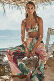 Woman in a colorful bikini and pants sitting under a thatched roof with a beach and mountains in the background.