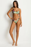 A woman modeling a two-piece bikini with a colorful abstract print in green, purple, orange, and white, set against a plain light gray studio background.