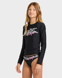 A young female model wearing a black long-sleeve Billabong rash guard top with a white and pink graphic print on the chest, paired with matching black swim briefs featuring a white and pink Billabong logo band.