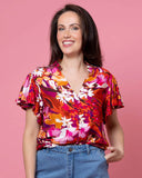 Woman wearing a colorful floral blouse and blue jeans against a pink background Charlo Alex Blouse - Orange Floral