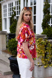 Woman wearing a colorful floral blouse and white shorts standing in front of a building with greenery. Charlo Alex Blouse - Orange Floral