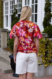Person wearing a colorful floral Charlo Alex Blouse - Orange Floral shirt and white shorts standing outdoors. 