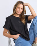 Woman wearing a black t-shirt with 'FOXWOOD' logo and blue jeans, sitting on a chair against a light gray background.
