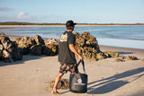 A man walks barefoot on a sandy beach with rocks and water in the background, carrying a black tote bag with white text that reads 'THE MAD HUEYS'. The Mad Hueys Parallel Hueys Dry Bucket Bag - Black