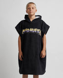 A child wearing a black hooded towel poncho with a large 'MAD HUEYS' graphic across the chest, standing against a light gray studio background.