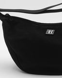 A close-up of a black nylon fanny pack with a curved shape, a top zipper, and a small white square logo patch on the front. The strap is visible at the top against a light gray background.