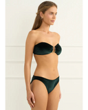 A woman modeling a dark green velvet two-piece bikini set against a clean white paneled wall.