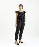 A woman modeling a black top with 4 tiers of frills and ruffled shoulders, paired with black pants.