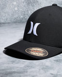 A black structured baseball cap with a white embroidered Hurley logo on the front and a gold and black Flexfit sticker on the brim, set against a gray textured background.