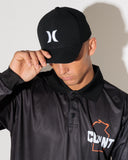 A man wearing a black Hurley baseball cap and a black Hurley polo shirt with a white logo on the front, posed against a light gray wall with a strong shadow.