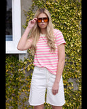 Woman wearing a pink and white striped shirt and white shorts, standing against a green ivy-covered wall.