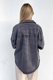 A woman viewed from behind wearing a faded charcoal gray washed denim shirt jacket and cream pants, standing against a plain white wall.