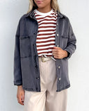 A woman wearing a charcoal gray button-up overshirt over a red and white striped t-shirt, paired with beige wide-leg trousers, standing against a white wall.