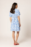 A woman is shown from the back wearing a short-sleeve blue and white patterned dress with a tie waist, paired with red heels, against a plain light beige studio background.