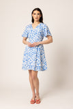 A woman modeling a short-sleeve V-neck dress with a blue and white tropical leaf pattern, paired with red strappy sandals, against a plain beige studio background.