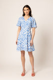A woman modeling a short-sleeve V-neck wrap dress with a blue and white leaf pattern, paired with red strappy sandals, against a plain beige studio background.
