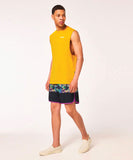 A man wearing a yellow sleeveless tank top and multicolored board shorts with a palm leaf pattern, standing against a plain background.