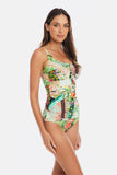 A woman modeling a one-piece swimsuit with a colorful tropical and scenic print featuring green foliage, blue water, and pink accents, set against a plain light gray background.