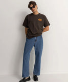 Person wearing a brown t-shirt with a logo and blue jeans against a white background Rhythm Essential Jean - Dark Blue
