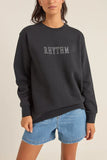 Person wearing a black sweatshirt with 'RHYTHM' text and blue shorts on a beige background