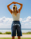 Woman standing with arms raised at the beach, wearing a striped tank top and denim shorts. Roxy logo on bum.