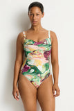 A woman modeling a one-piece swimsuit with a colorful abstract swirl pattern in green, pink, yellow, and white against a plain light gray studio background.