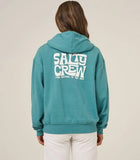 A woman viewed from the back wearing a teal hoodie with a large white 'SALTY CREW' graphic on the back, paired with white pants, against a plain beige studio background. Salty Crew Womens Freestyle Hoodie - Teal