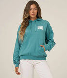 A woman wearing a teal ribbed pullover hoodie with a white 'SALTY CREW' graphic on the chest, paired with white pants, against a plain light beige studio background. Salty Crew Womens Freestyle Hoodie - Teal
