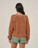 A woman shown from the back wearing a rust-colored, open-knit long-sleeve sweater and sage green shorts against a plain light beige studio background. Back view of the a woman wearing the Salty Crew Womens Surge Sweater - Mocha with green shorts on a white background.