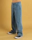 A person wearing light blue wide-leg denim jeans and black low-top sneakers, standing against a solid mustard yellow studio background. Santa Cruz Yelling Stack Baggy Denim Jeans - Mid Blue Denim