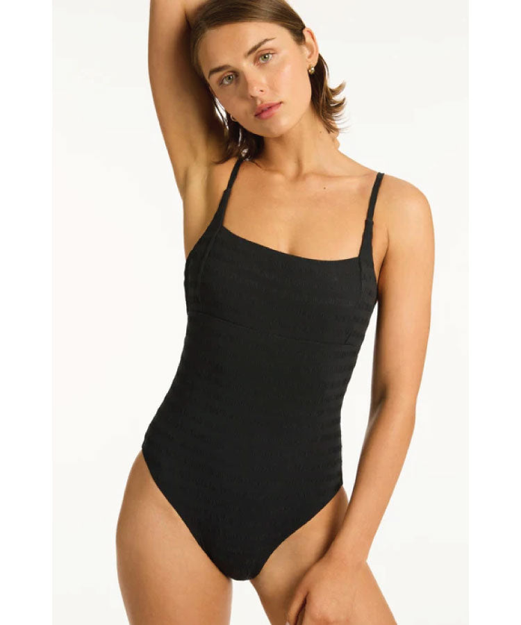 A model wearing a solid black square neck bralette one-piece swimsuit with adjustable straps.
