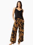 A woman modeling a two-piece loungewear set featuring a black camisole top with lace trim and wide-leg pants with a black and mustard yellow baroque pattern, set against a plain white background. Woman wearing the Seeking Lola Every Day Love Pants - Burnt Sienna