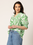 A woman modeling a green and white abstract patterned blouse with puff sleeves and a split neckline, paired with light blue jeans, against a plain light beige studio background. Seeking Lola SS Gifted Top - Green Mirage