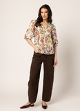 A woman modeling a short-sleeved floral blouse with brown wide-leg pants and dark brown flats, standing against a plain beige studio background. Seeking Lola Graceful Top - Ballet Blossom