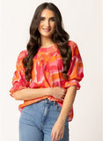 A woman modeling a vibrant, short-sleeved blouse with a wavy abstract pattern in shades of orange, pink, and red, paired with blue jeans against a plain white studio background.