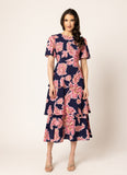 A woman modeling a short-sleeve midi dress with a navy blue base and a pink floral print, standing against a plain light beige studio background.