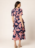 A woman shown from the back wearing a navy midi dress with a pink floral print, short sleeves, and a tiered ruffle hem, paired with beige block-heel sandals against a plain light beige studio background.