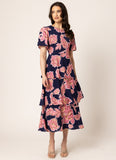 A woman modeling a navy blue midi dress with a pink floral print, featuring short sleeves, a round neckline, and a tiered skirt, set against a plain beige studio background.