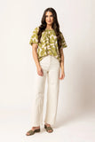 A woman modeling a short-sleeve blouse with an olive green and cream leaf pattern, paired with cream wide-leg pants and olive green slide sandals, against a plain off-white studio background.