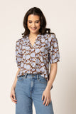 A woman modeling a short-sleeve button-up blouse with a brown, white, and blue floral pattern, paired with light-wash blue jeans, against a solid light beige background.