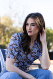 A woman wearing a brown and blue floral patterned short-sleeve blouse and blue jeans, sitting outdoors on a striped cushion.