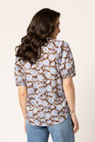 A woman shown from the back wearing a short-sleeve button-up shirt with an all-over brown, beige, and blue floral pattern, paired with blue jeans, against a plain off-white background.