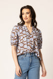 A woman modeling a short-sleeve button-up blouse with a brown, white, and light blue floral pattern, paired with light-wash blue jeans, against a solid off-white background.