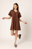 A woman modeling a brown short-sleeve V-neck tunic dress with a scalloped hem, posed against a plain light beige studio background.