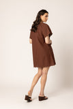 A woman modeling a brown short-sleeve dress with a scalloped hem, shown from a side-back angle against a plain light beige studio background. Seeking Lola Scallop Shortie Dress - Chestnut