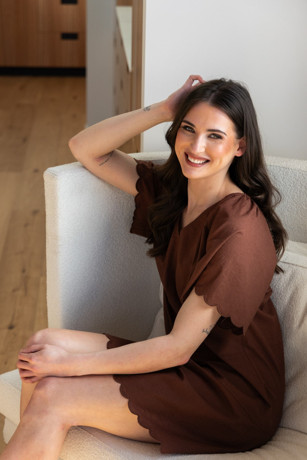 A woman with long dark hair sits smiling on a light beige upholstered chair in a bright indoor setting. She is wearing a brown short-sleeve dress with scalloped edges. Seeking Lola Scallop Shortie Dress - Chestnut