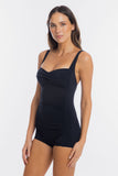 A woman modeling a black ruched one-piece swimsuit with shoulder straps and mid-thigh shorts against a plain light gray studio background.